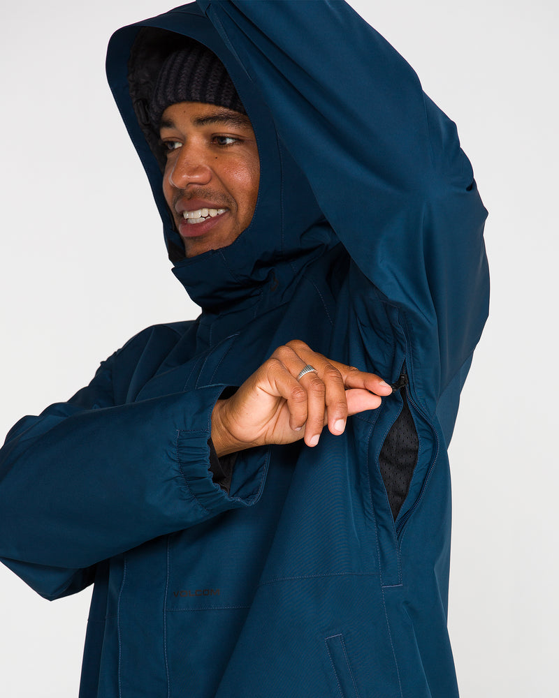 Wearing the Volcom Men’s Dua Gore-Tex Jacket in blue and a black beanie, a person unzips an underarm vent, smiling confidently as the GORE-TEX waterproof protection keeps them dry and comfortable.