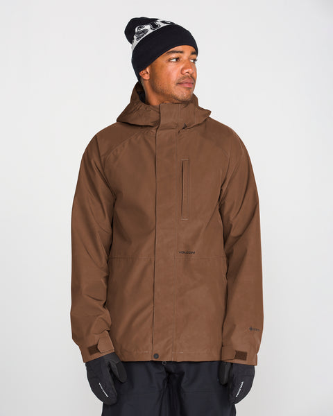 A person wearing the Volcom Men’s Dua Gore-Tex Jacket in brown, paired with black gloves, black pants, and a black beanie featuring a white pattern, stands against a plain white background looking slightly to the side. The jacket delivers dependable GORE-TEX waterproof protection while blending style with functionality.