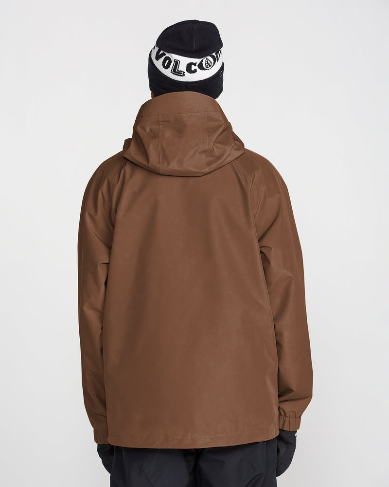 A person stands with their back to the camera against a plain white background, wearing the Volcom Men’s Dua Gore-Tex Jacket in brown for waterproof protection, along with black pants, black gloves, and a black beanie featuring white VOLCOM text.
