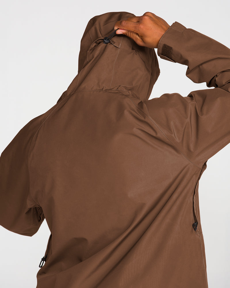 A person wearing the Volcom Men’s Dua Gore-Tex Jacket in brown is seen from behind, adjusting the hood with their right hand against a plain white background. This GORE-TEX jacket from Volcom provides reliable waterproof protection for any adventure.