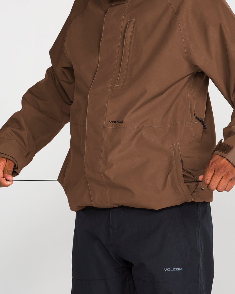 A person wearing a brown Volcom Men’s Dua GORE-TEX Jacket and black Volcom pants adjusts the drawstring at the hem of the jacket. The image centers on their torso and hands, emphasizing the jacket’s premium GORE-TEX waterproof protection, while the face remains out of view.