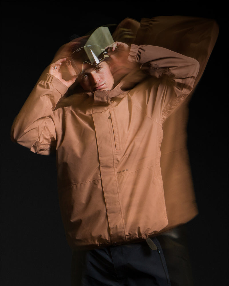 Wearing the Volcom Men’s Dua Gore-Tex Jacket and dark ski pants, a person adjusts their visor, captured with a dynamic motion blur effect against a black background. The outfit’s waterproof construction highlights its suitability for active conditions.