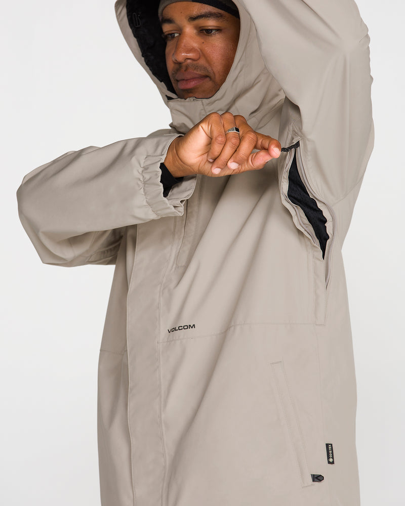 A person wearing the Volcom Men’s Dua Gore-Tex Jacket in beige, featuring GORE-TEX waterproof protection, uses one hand to unzip a black underarm vent. The jacket displays a subtle Volcom logo near the chest and has a zippered pocket on the front. The background is plain white.
