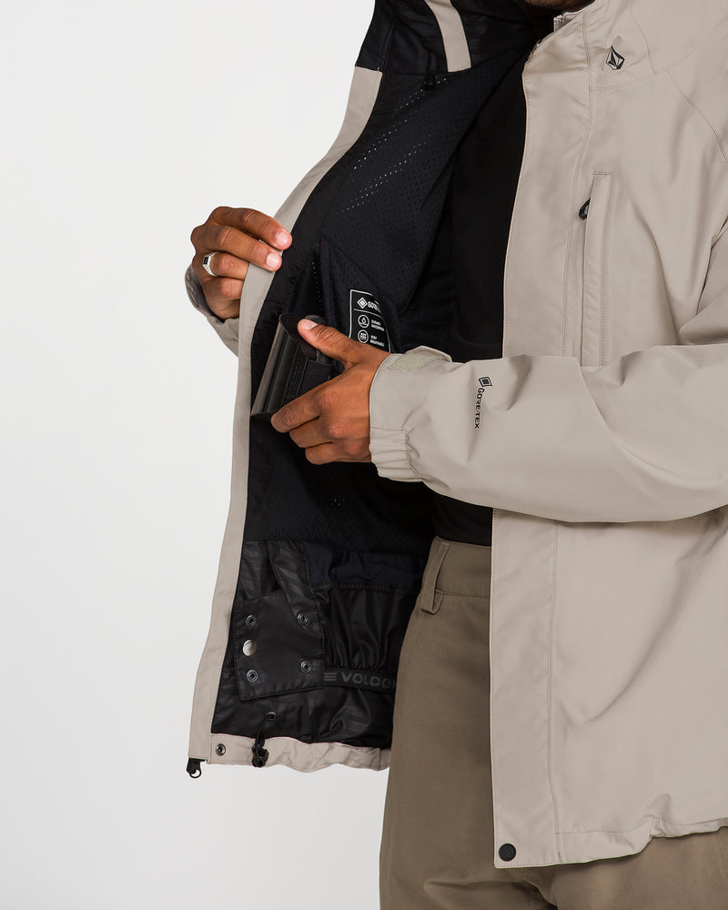A person wearing the Volcom Men’s Dua Gore-Tex Jacket in beige holds it open, revealing an interior zippered pocket and mesh lining designed for waterproof protection. Only the torso and hands are visible against a plain white background.