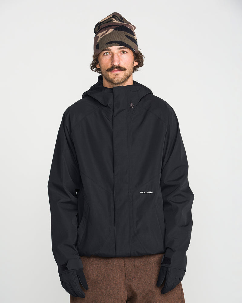 A man with a mustache stands against a plain white background wearing the Volcom Men’s Collins 20K Jacket—a black waterproof hooded jacket made from a recycled shell—paired with brown pants, black gloves, and a brown camo beanie.