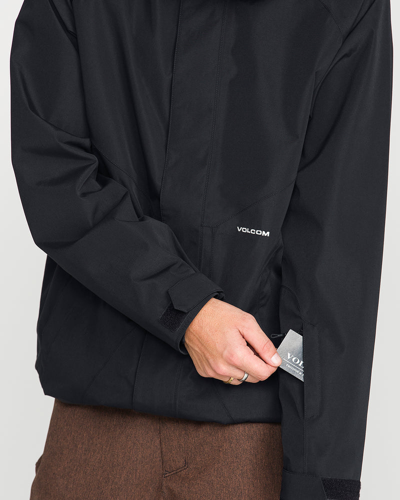 Wearing a black Volcom Men’s Collins 20K Jacket and brown pants, a person pulls out a gray VOLCOM tag from a small zippered pocket on the left sleeve.