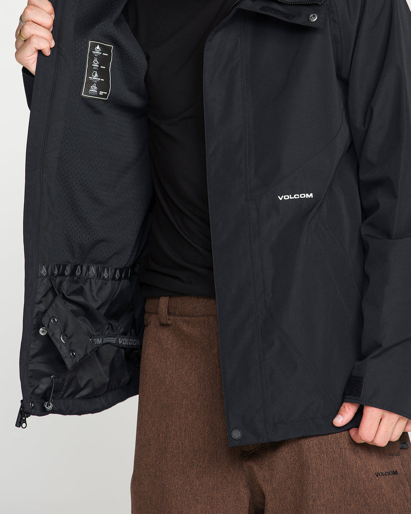 A person wearing brown pants and a black Volcom Men’s Collins 20K Jacket opens the jacket to reveal its inner lining and labels. The Volcom logo is visible on the outside of this waterproof jacket.