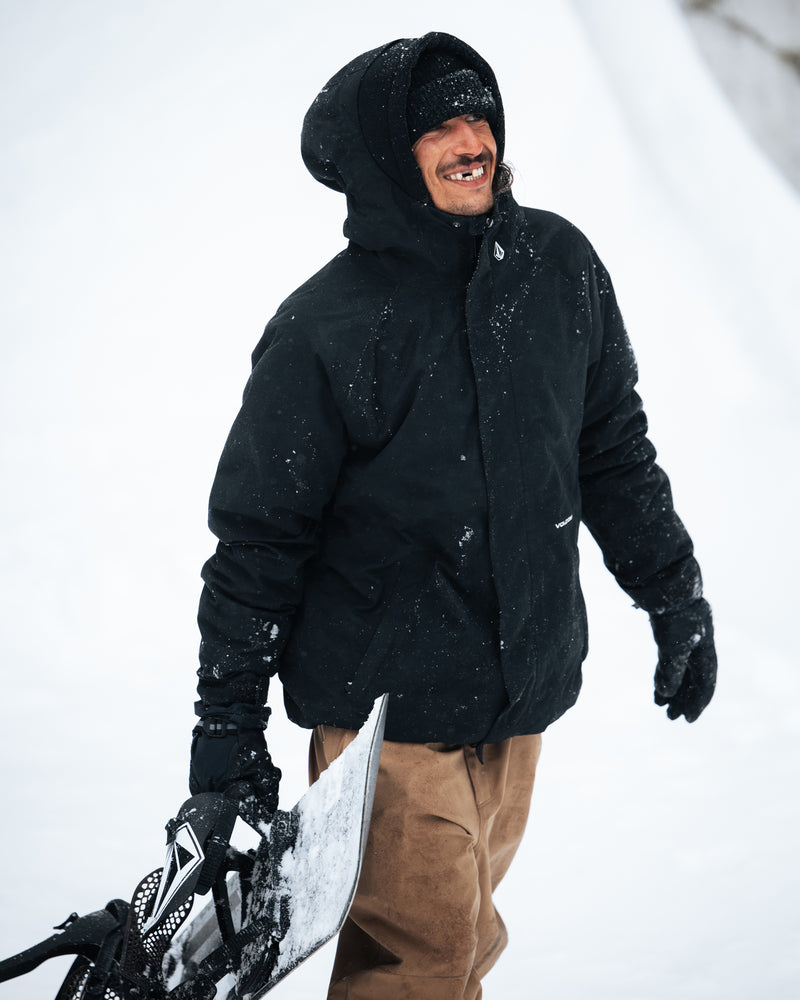 A person in the Volcom Men’s Collins 20K Jacket with a recycled shell and tan pants smiles while holding a snowboard in a snowy outdoor setting.