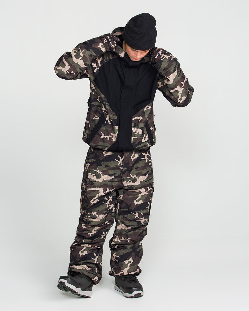 A person wearing a black beanie, Volcom Men’s Collins 20K Jacket with a camo-patterned design, and matching pants stands against a plain white background, looking down and adjusting their jacket.