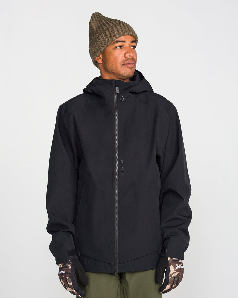 A person wearing the Volcom Men’s Brighton Full Zip Jacket in black—crafted from V-Science 2-Layer shell with 15K/15K protection—paired with a green knit beanie, camouflage gloves, and olive pants stands against a plain white background, looking slightly to the side.