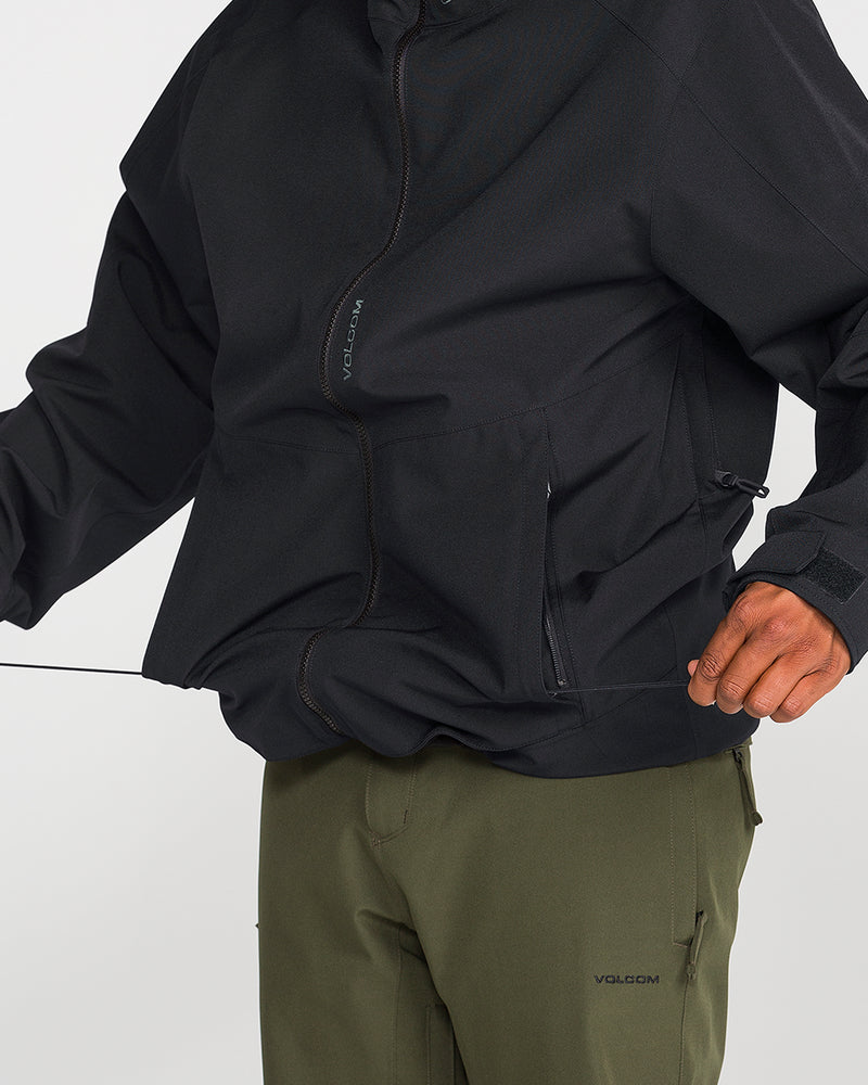 A person wearing the Volcom Men’s Brighton Full Zip Jacket in black, made from V-Science 2-Layer shell with zippered pockets and 15K/15K protection, adjusts the jacket’s drawstring. The Volcom logo is visible on their green pants.