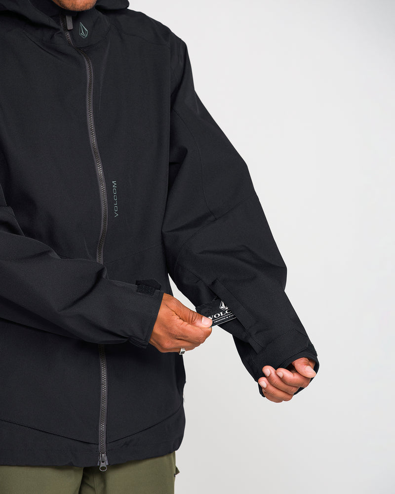 A person wearing the Volcom Men’s Brighton Full Zip Jacket, which features a V-Science 2-Layer shell, adjusts a strap on the sleeve. Only the torso and arms are visible, and they are also wearing olive-green pants.