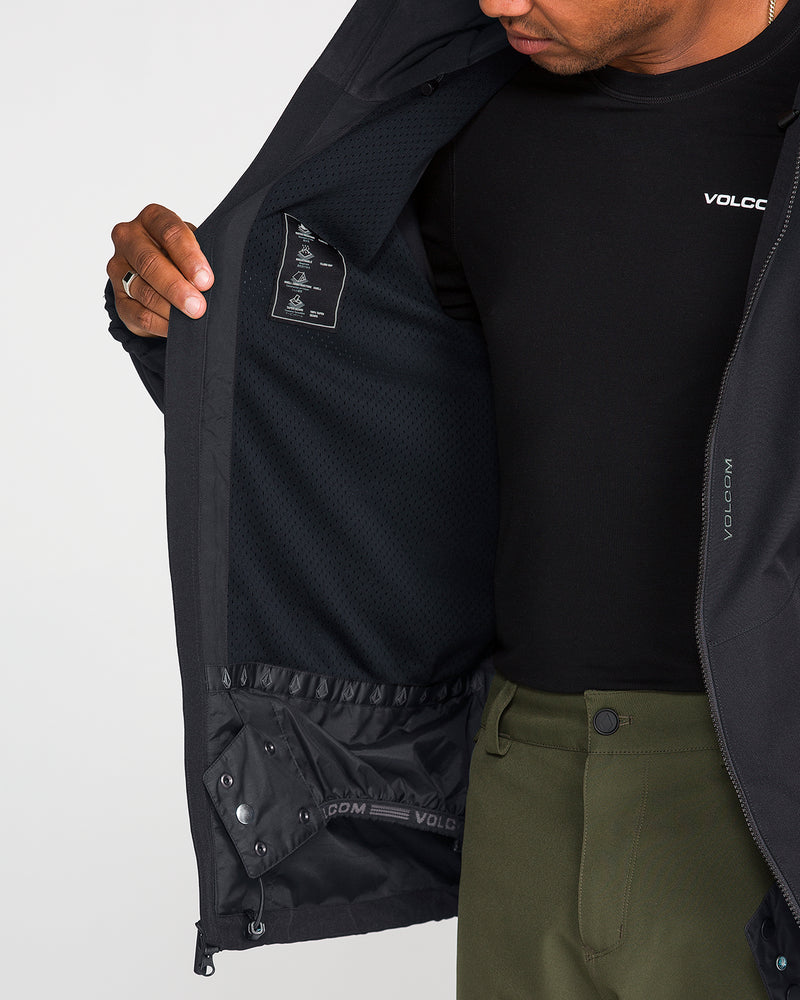 Wearing a black Volcom Men’s Brighton Full Zip Jacket, a person opens it to reveal the mesh lining and inner details, including the advanced V-Science 2-Layer shell. They are also dressed in olive green pants and a black Volcom shirt, with their face partially out of frame.