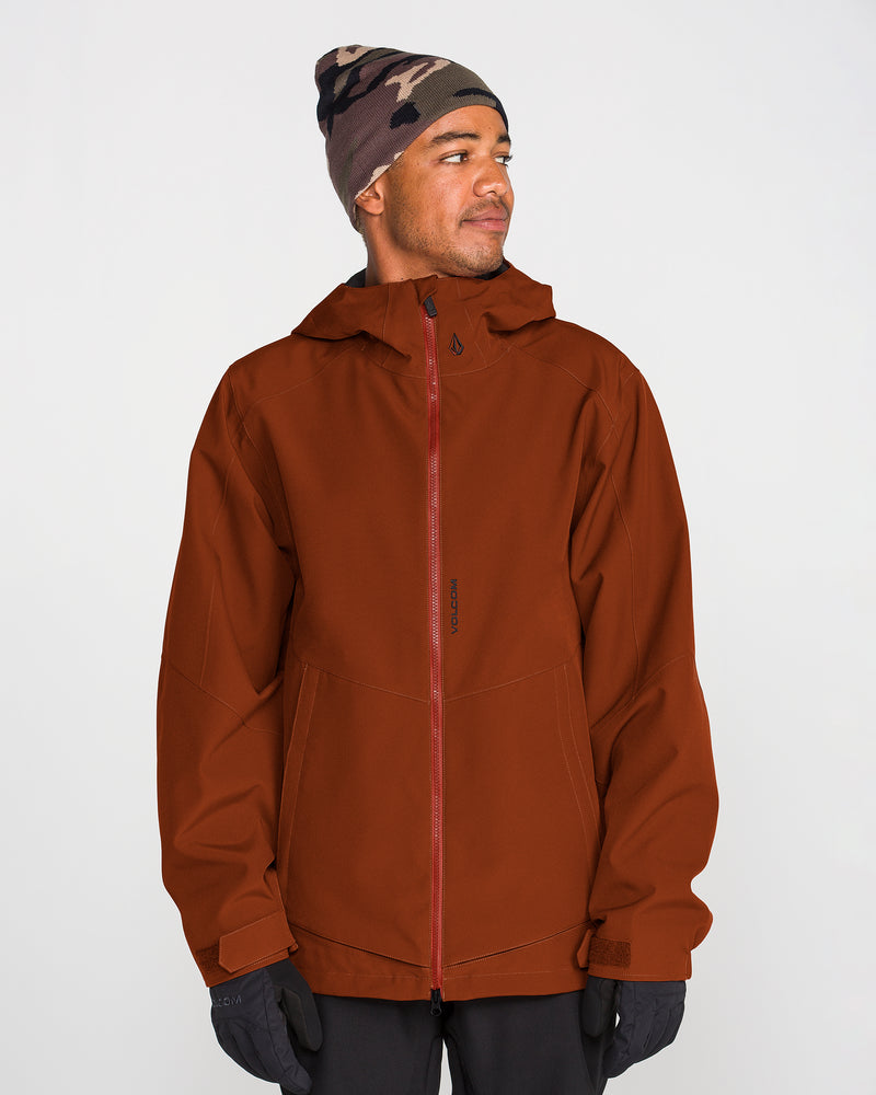 A person wearing the Volcom Men’s Brighton Full Zip Jacket in rust, featuring a V-Science 2-Layer construction and PFAS-free DWR, along with black gloves and a patterned gray beanie, stands turned slightly to the side and looks off-camera against a plain light background.