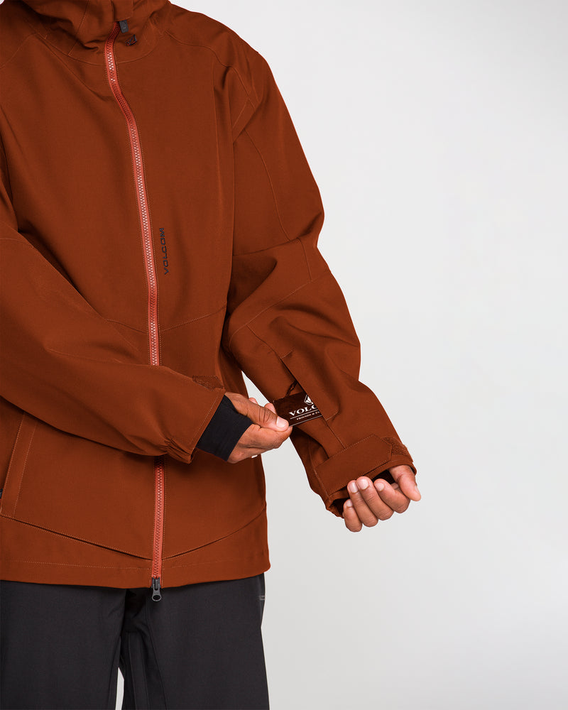 A person wearing the Volcom Men’s Brighton Full Zip Jacket in a rust color unzips a small pocket on the left sleeve, revealing a card partially inside. The jacket features a red zipper, PFAS-free DWR coating for added protection, and black pants are visible.