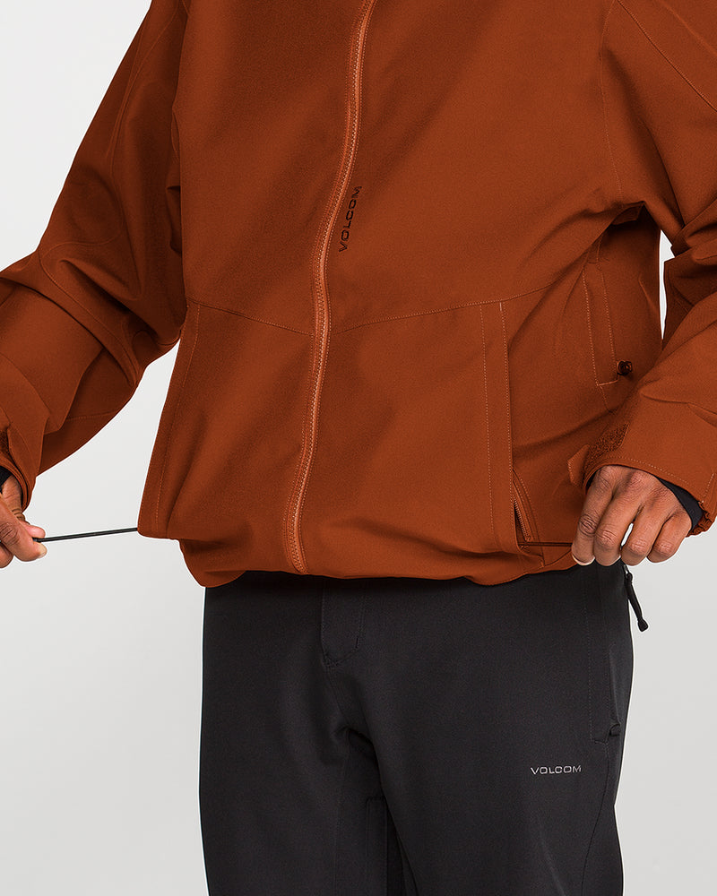 A person wearing Volcom’s Men’s Brighton Full Zip Jacket in rust and black pants adjusts the drawstring at the jacket’s hem; only their torso and hands are visible.