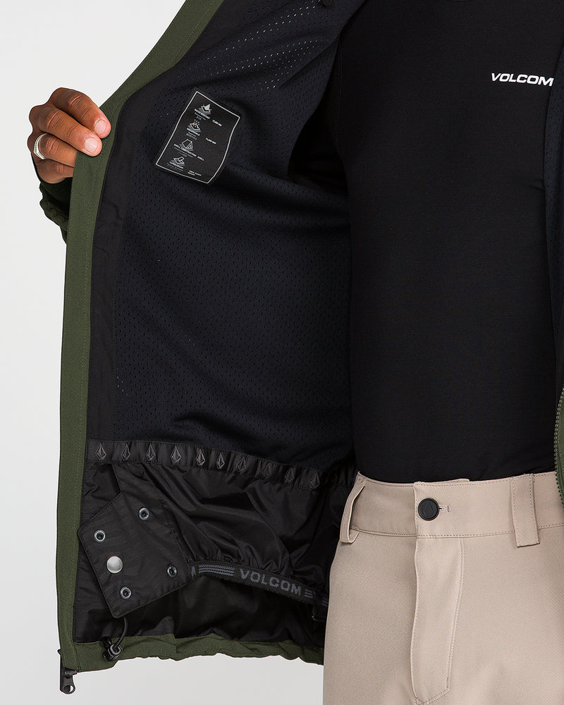 A person opens a green Volcom Men’s Brighton Full Zip Jacket to reveal its black mesh inner lining and interior tag, while wearing beige pants and a black VOLCOM shirt.