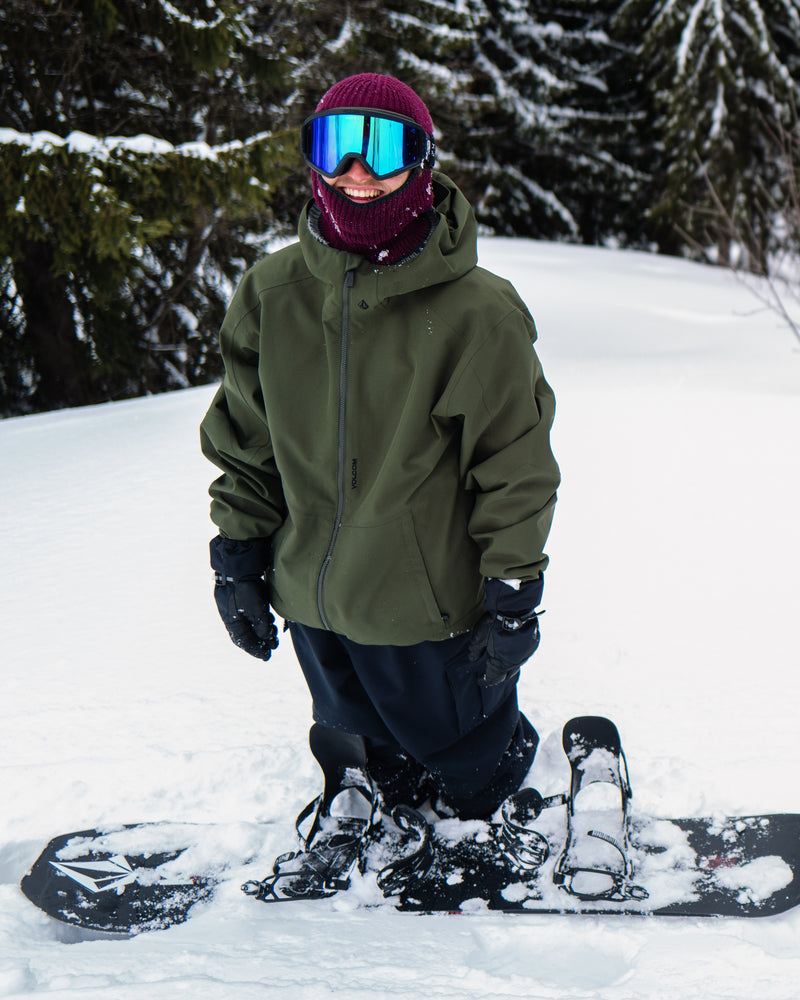 A person in winter gear, wearing a green Volcom Men’s Brighton Full Zip Jacket and reflective goggles, kneels in the snow next to a snowboard with a forest in the background.