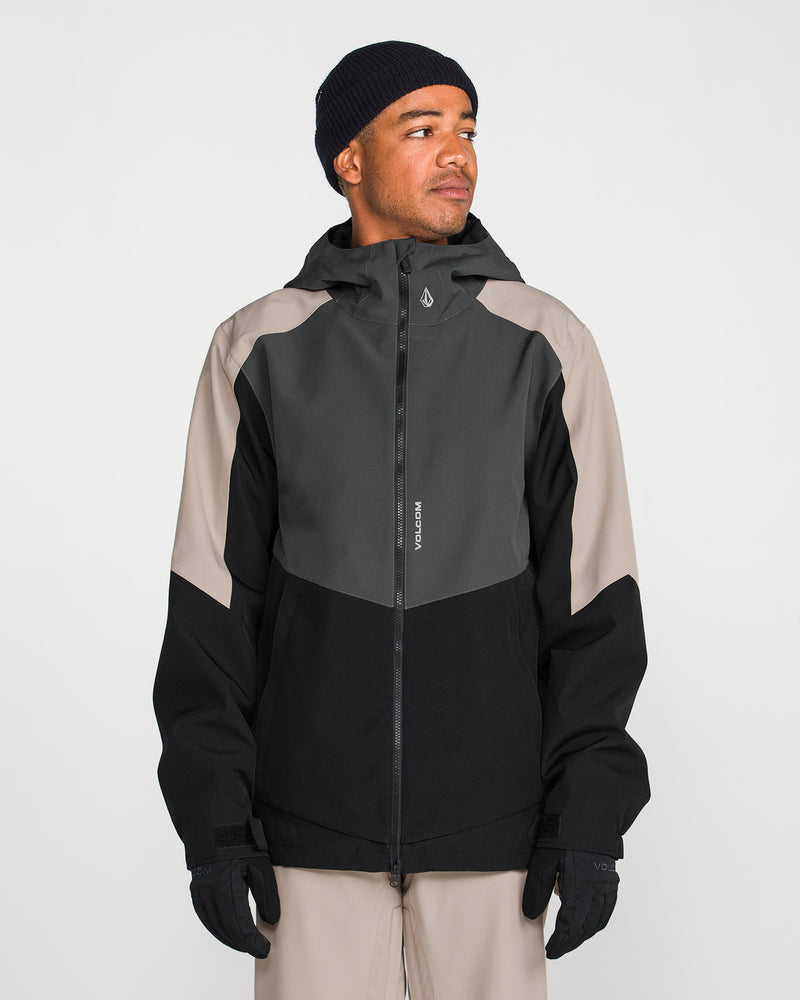 A person wearing the Volcom Men’s Brighton Full Zip Jacket—a black and gray hooded jacket with V-Science 2-Layer construction and a PFAS-free DWR finish—along with beige pants, black gloves, and a black beanie, stands in front of a plain white background, looking slightly to the side.