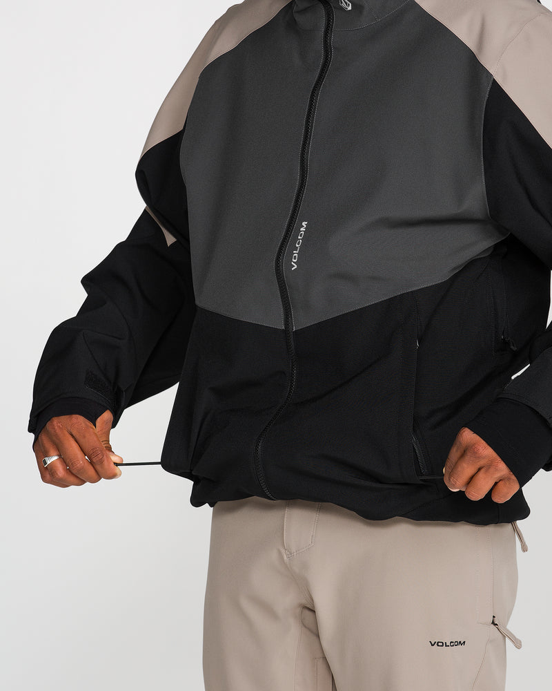 A person wearing the Volcom Men’s Brighton Full Zip Jacket with bold color-blocking and beige pants adjusts the drawstring at the hem. The jacket, displaying “VOLCOM” on both the zipper and chest, combines style and performance with a PFAS-free DWR finish. The person’s head is not visible.