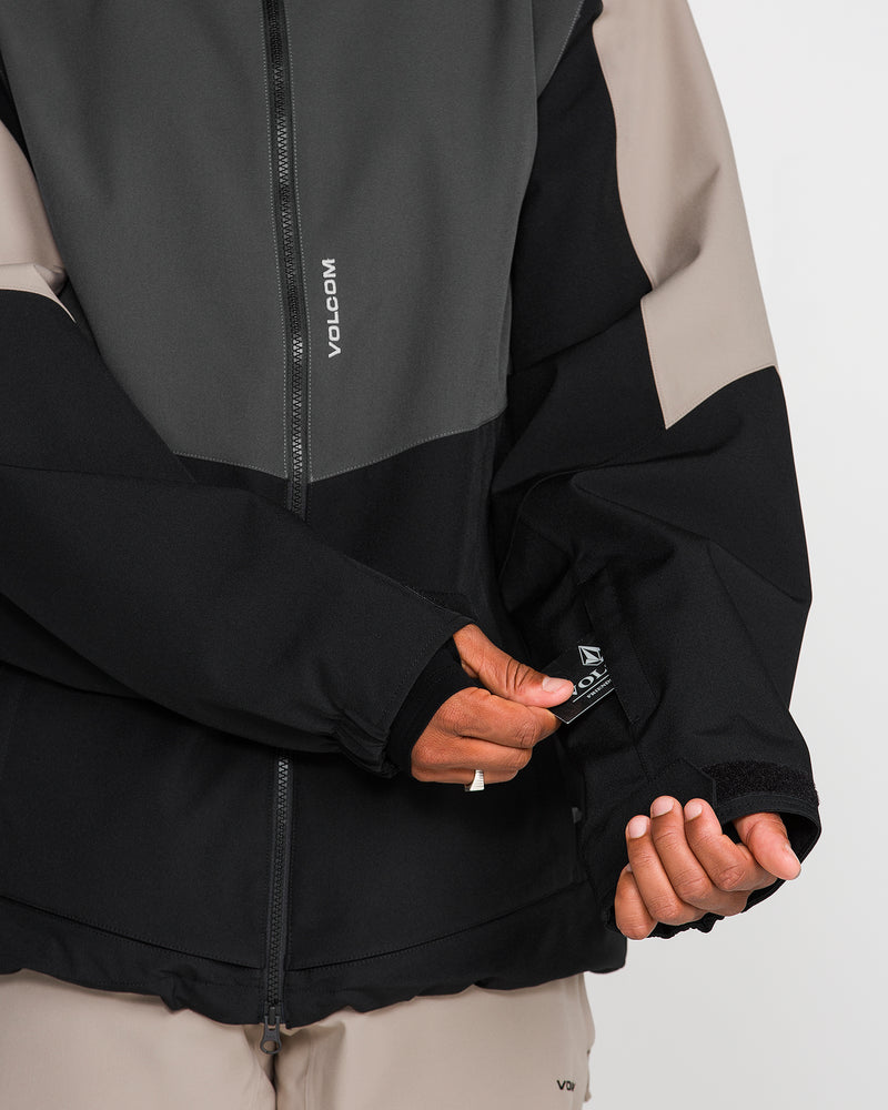 A person wearing the Volcom Men’s Brighton Full Zip Jacket, which features a color-block design in black, gray, and beige, adjusts the Velcro strap on the sleeve. The jacket displays VOLCOM branding on both the zipper and sleeve tag.
