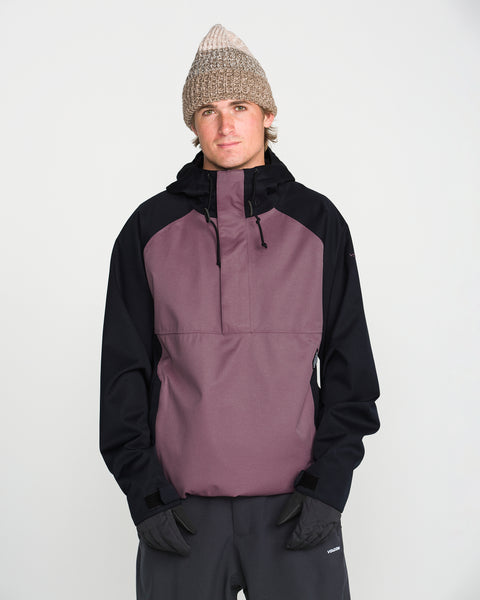 A person is pictured against a plain white background, wearing a beige knit beanie, black gloves, dark pants, and the Volcom Men’s Hossegor 20K Pullover—a purple and black jacket made with a PFAS-free DWR recycled V-Science shell—looking directly at the camera with their hands in their pockets.