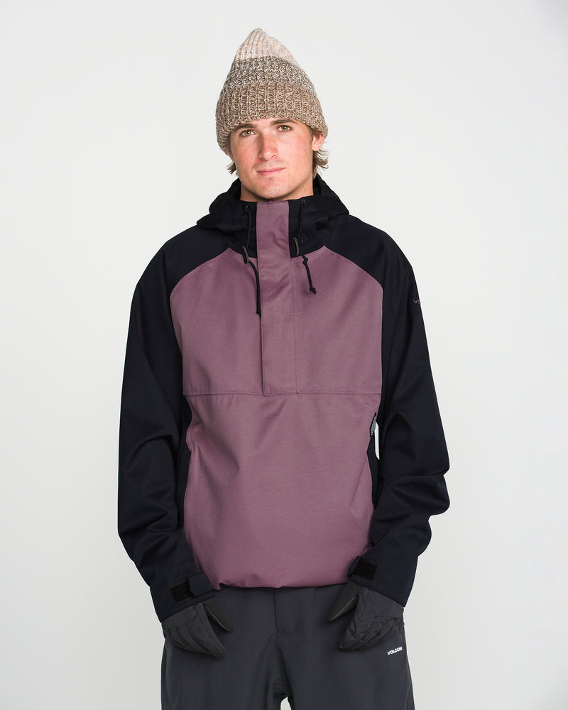 A person is pictured against a plain white background, wearing a beige knit beanie, black gloves, dark pants, and the Volcom Men’s Hossegor 20K Pullover—a purple and black jacket made with a PFAS-free DWR recycled V-Science shell—looking directly at the camera with their hands in their pockets.