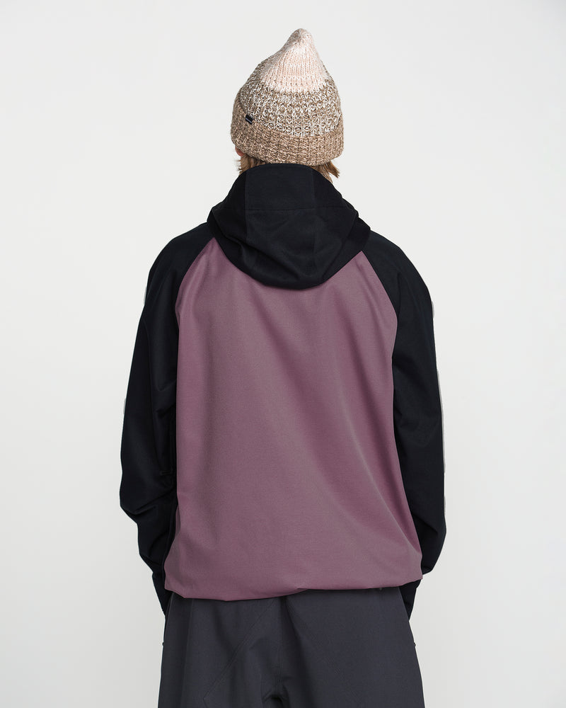 A person wearing the Volcom Men’s Hossegor 20K Pullover—a two-tone jacket with black sleeves and a mauve back panel, crafted from recycled V-Science shell—and a brown knit beanie, stands with their back to the camera against a plain white background.