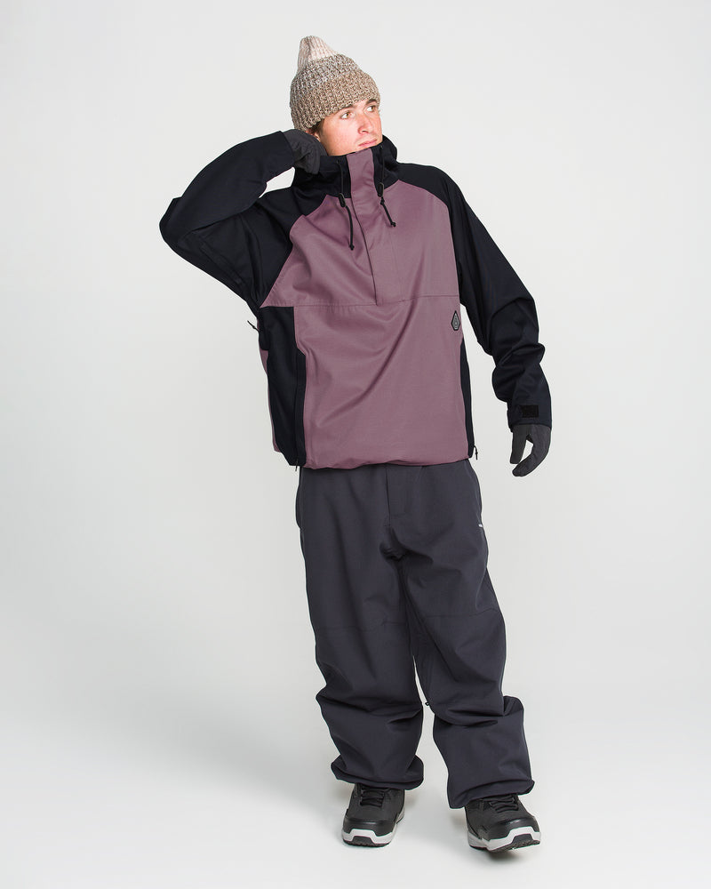 A person dressed for winter, featuring the Volcom Men’s Hossegor 20K Pullover—a color-blocked jacket crafted from a recycled V-Science shell with PFAS-free DWR—along with a gray knit hat, dark pants, gloves, and sneakers, stands on a white background with one arm bent and touching their head.