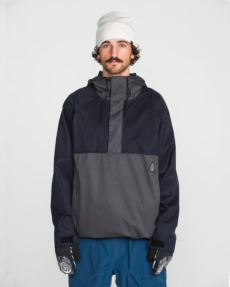 A man with a mustache, wearing a white beanie, blue pants, and patterned gloves, stands facing forward against a plain white background. He is dressed in the Volcom Men’s Hossegor 20K Pullover jacket, crafted from recycled V-Science shell in black and gray.