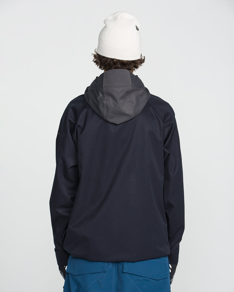 A person wearing a white beanie and blue pants is standing facing away from the camera against a plain light background, dressed in the Volcom Men’s Hossegor 20K Pullover—a dark hooded jacket crafted from a recycled V-Science shell.