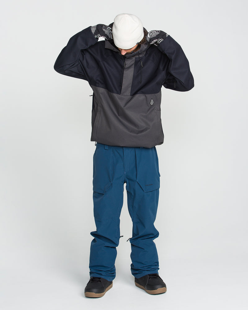 A person wearing the Volcom Men’s Hossegor 20K Pullover, a dark jacket crafted from recycled V-Science shell, along with blue pants, black shoes, patterned gloves, and a white beanie looks down while adjusting their hood against a plain white background.