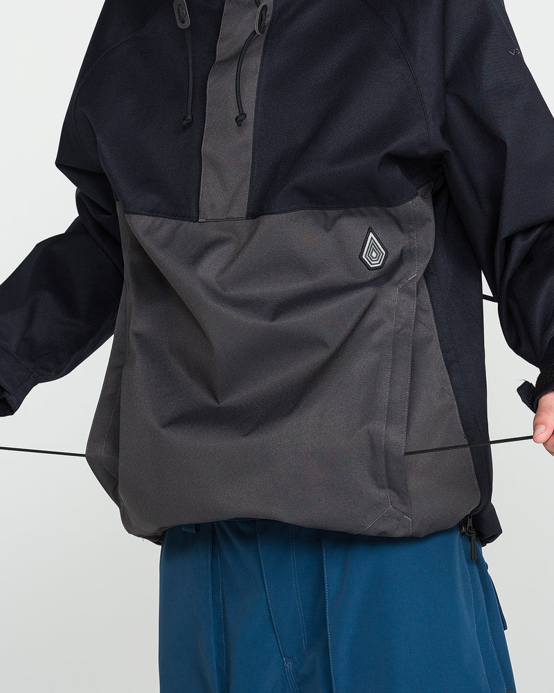 A person wearing the Volcom Men’s Hossegor 20K Pullover, featuring a front pocket and recycled V-Science shell construction, is adjusting a drawstring. The dark jacket is paired with blue pants, with the torso and part of the arms visible.