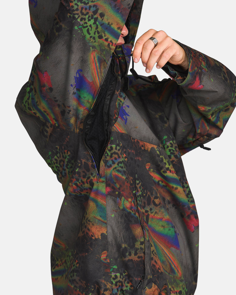 A person wearing the Volcom Men’s Hossegor 20K Pullover, a colorful, patterned jacket with animal print details, is unzipping a side zipper near the armpit area. The hood is up, partially obscuring their face. This pullover features a recycled V-Science shell for sustainable style and performance.