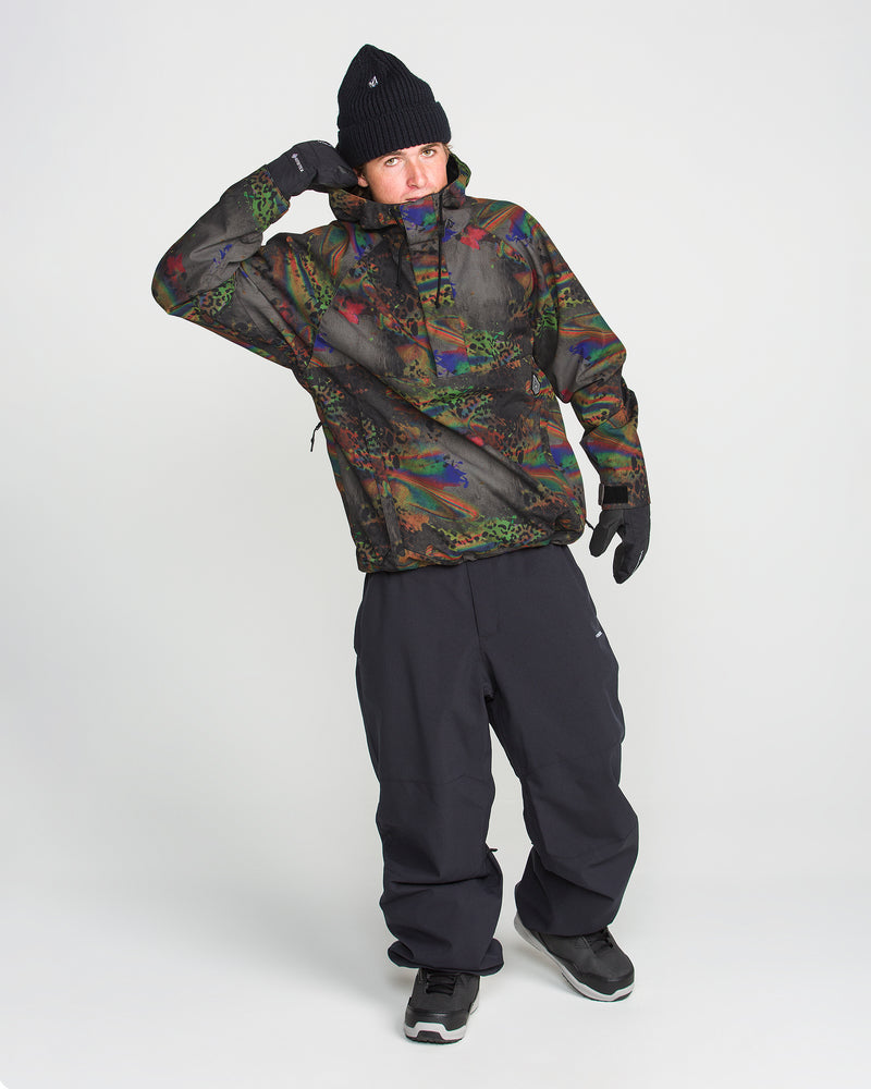 A person in the Volcom Men’s Hossegor 20K Pullover, featuring a colorful camouflage recycled V-Science shell, black pants, gloves, sneakers, and a knit hat stands on a white background, raising one gloved hand to their head.