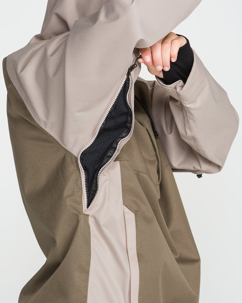 A person pulls up the side zipper of the Volcom Men’s Hossegor 20K Pullover—a two-tone beige and olive jacket with a PFAS-free DWR finish—revealing its black inner lining. Only the torso and part of an arm are visible.