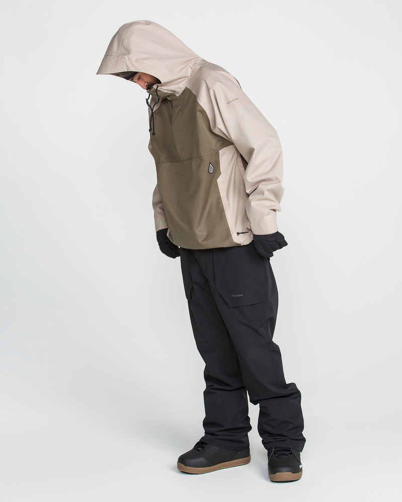 A person wearing the Volcom Men’s Hossegor 20K Pullover in beige—crafted from a recycled V-Science shell with PFAS-free DWR—stands looking down, paired with black gloves, black pants, and brown shoes against a plain white background.