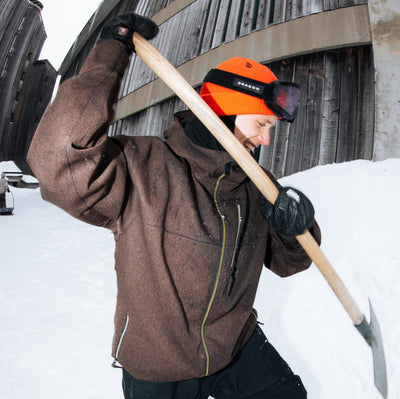 A person in a snowy outdoor setting wearing a brown technical snow jacket, an orange winter hat with goggles, and black gloves, holding a shovel.