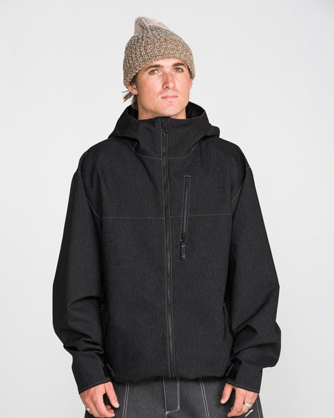 A person wearing the Volcom Men’s Top Stitch Jacket in black, styled with a baggy fit, and a knitted beige beanie stands against a plain light background, gazing slightly upward with a neutral expression.