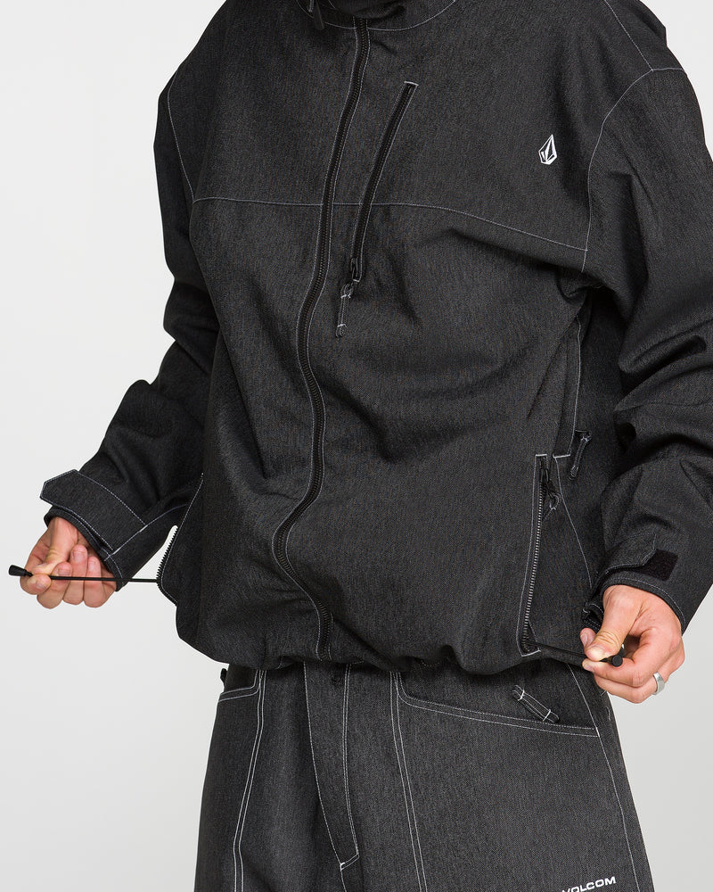A person is seen wearing the Volcom Men’s Top Stitch Jacket in dark gray with matching pants, holding the jacket’s drawstring at the hem. This baggy fit jacket showcases white stitching and a small logo on the chest. The person’s face is not visible.