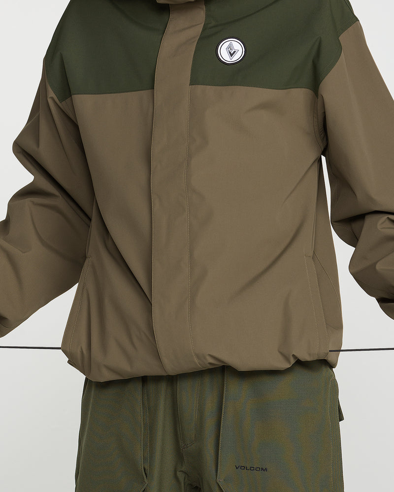 A person is wearing the Volcom Men’s Buckthreeeighty Jacket in olive green and brown, featuring a chest patch and a baggy fit. The V-Science 2-Layer jacket offers added protection with PFAS-free DWR, paired with matching pants. The individual’s arms are slightly raised, and their head is not visible in the image.