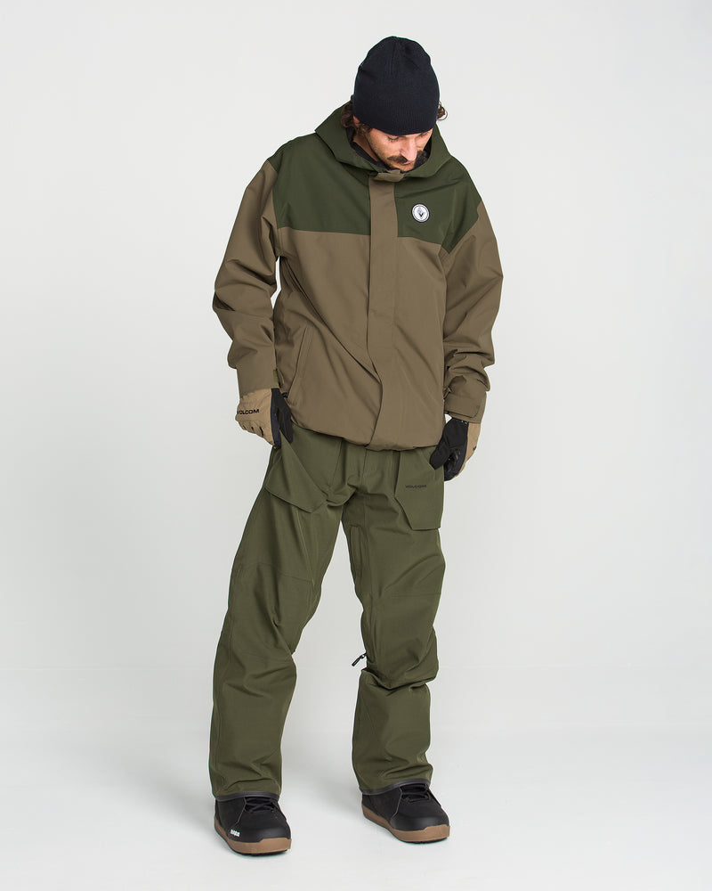 A person wearing the Volcom Men’s Buckthreeeighty Jacket in an olive green and brown baggy fit, along with matching pants, a black beanie, black gloves, and black shoes stands against a plain white background, looking downward. The jacket features V-Science 2-Layer construction and a PFAS-free DWR finish for comfort and eco-friendly protection.