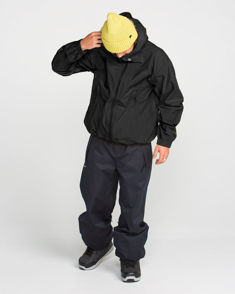 A person in a yellow beanie and black hooded jacket—specifically the Volcom Men’s Arthur 3L 20K Jacket—paired with dark pants and black boots stands looking down, one hand touching their head, against a plain white background.
