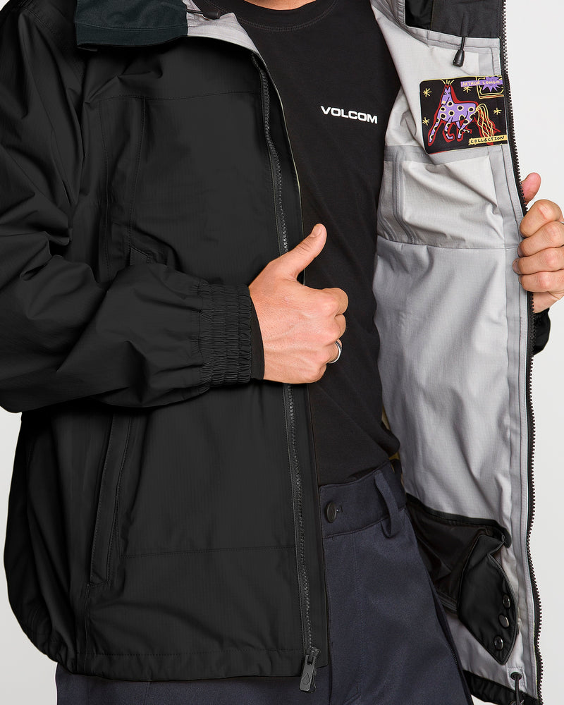 A person wearing the Volcom Men’s Arthur 3L 20K Jacket in black holds it open to display the gray interior lining and a vibrant inner patch with an illustrated alien and stars. Constructed with storm-ready V-Science 3-Layer protection, only their torso is visible, and they wear a black shirt underneath.