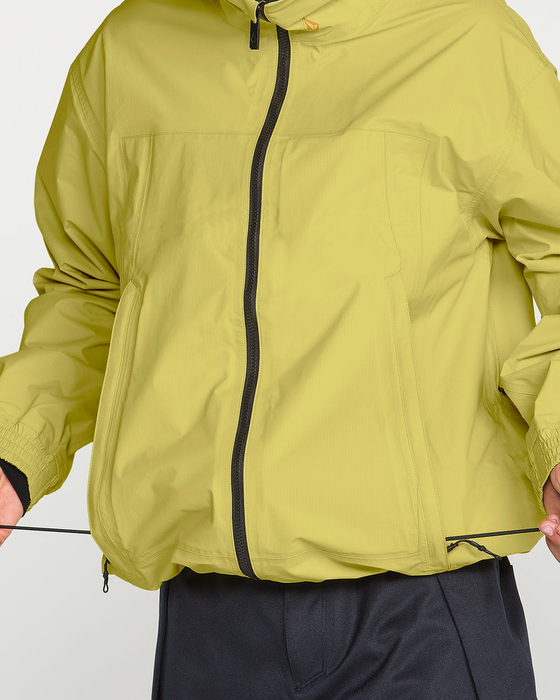 A person wearing a light green Volcom Men’s Arthur 3L 20K Jacket is adjusting the jacket’s waist drawstring. Only the torso and part of the arms are visible.