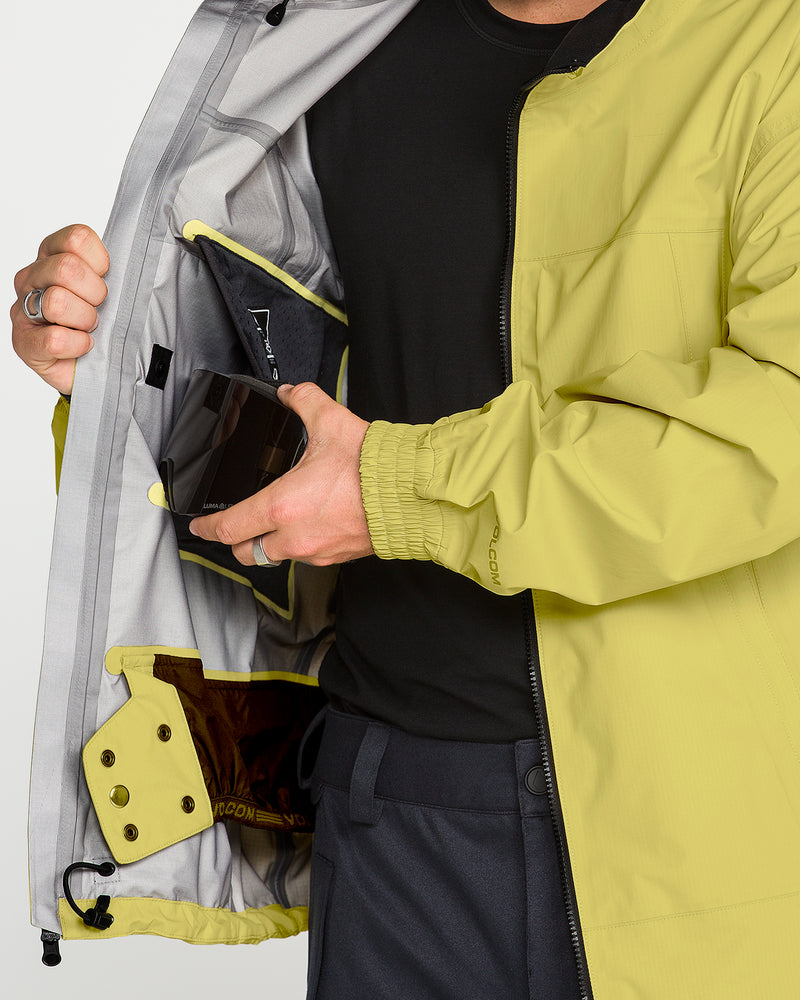 A person in a yellow Men’s Arthur 3L 20K Jacket by Volcom holds a phone, demonstrating the jacket’s phone pocket and interior features. The V-Science 3-Layer construction, gray lining, and extra small pockets are visible for added convenience. With only the lower half of the person’s face and body shown, the storm-grade protection and practical design of this Volcom jacket are emphasized.