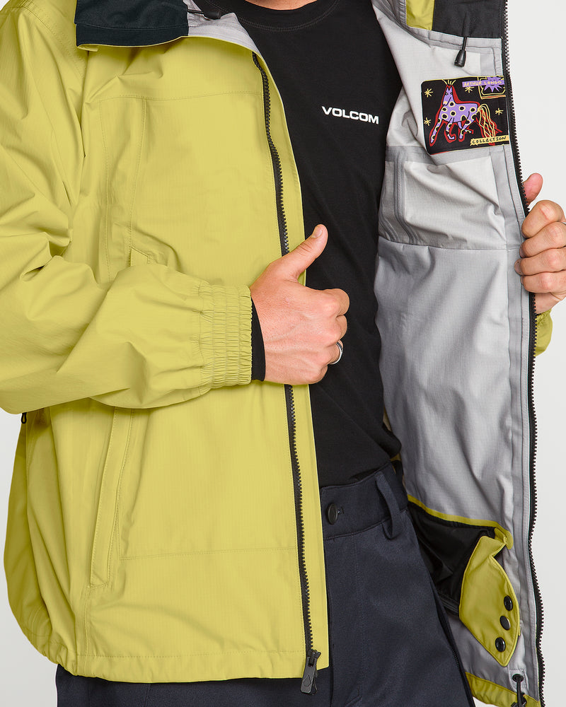 A person in a lime green Volcom Men’s Arthur 3L 20K Jacket holds it open to reveal the gray inner lining and an embroidered graphic patch labeled “APPROVED” with colorful designs near the inside pocket, showcasing its V-Science 3-Layer construction for storm-grade protection.