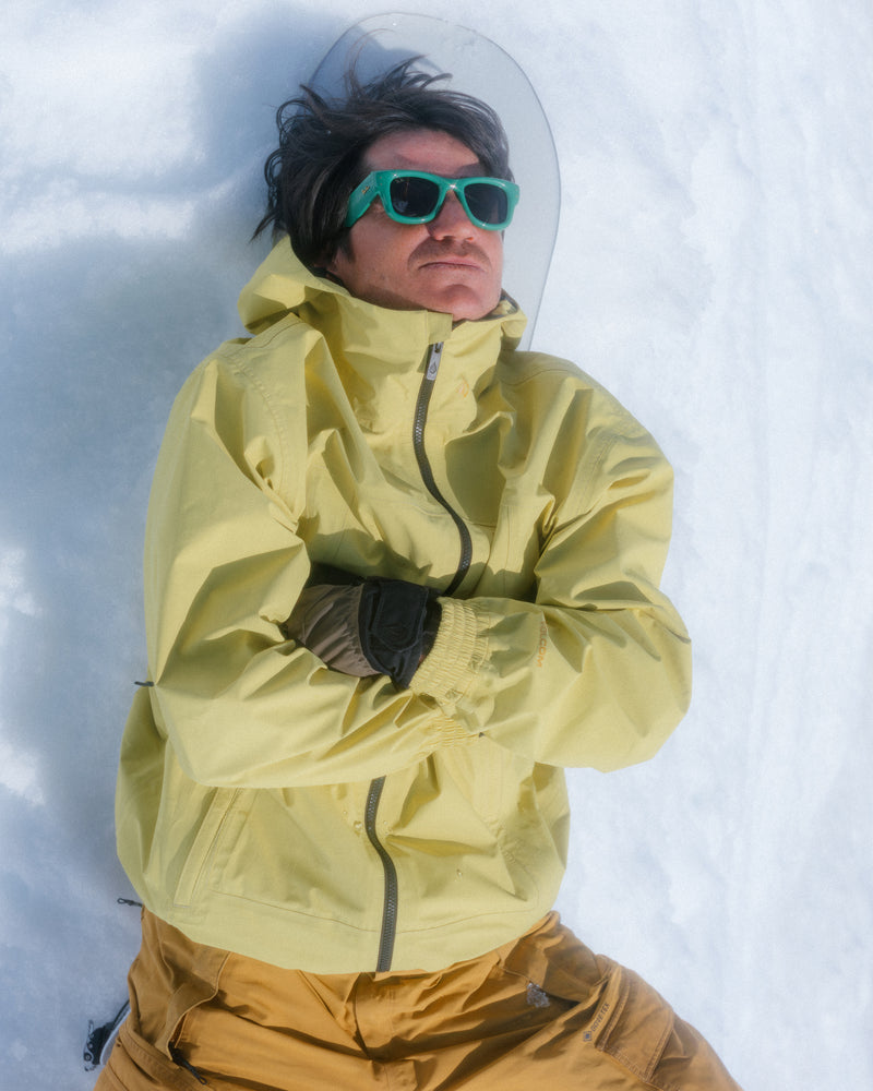 Clad in a yellow Volcom Men’s Arthur 3L 20K Jacket and green sunglasses, a person lies on their back in the snow with arms crossed and a snowboard peeking out behind their head—ready for anything with storm-grade protection.