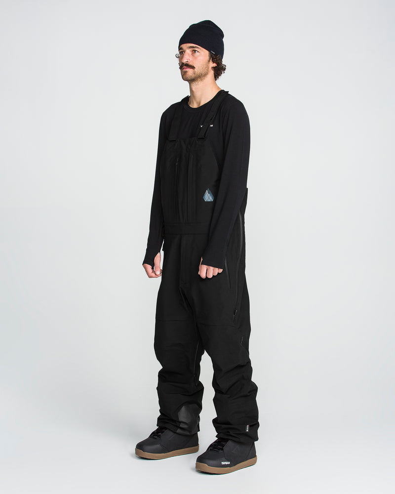 A person with a mustache and curly hair stands facing slightly left against a plain white background. They are wearing a black beanie, a long-sleeve shirt, large black boots, and the Volcom Men’s Anyox 3L Gore Pro Bib—with fully taped seams.
