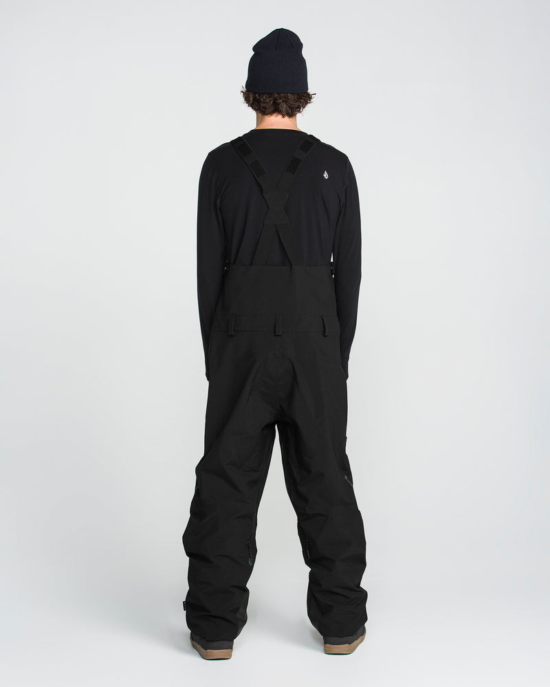 A person stands facing away from the camera against a plain white background, wearing a black beanie, long-sleeve shirt, and the Volcom Men’s Anyox 3L Gore Pro Bib—crafted with fully taped seams.